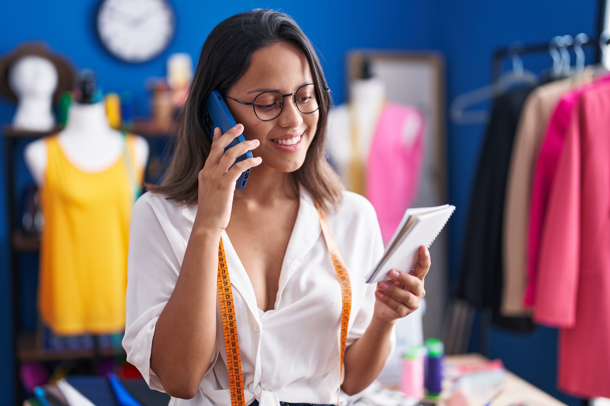 Young hispanic woman tailor talking on smartphone reading notebook at sewing studio